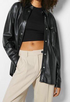 Frau trägt ein schwarzes bauchfreies Oberteil, eine offene schwarze Lederjacke und beige, hoch taillierte, plissierte Hosen, die Hände in den Taschen.