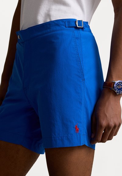 Blå shorts laget av lett materiale, med flat front, beltehempe og rødt brodert logo på kanten.
