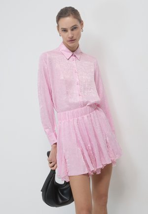 Camicia rosa trasparente con bottoni, maniche lunghe, tessuto testurizzato e vita arricciata, abbinata a una gonna a campana coordinata. Borsa nera.