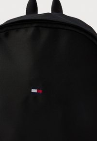 Črn nahrbtnik iz tkanine z teksturirano površino; na spodnjem levem vogalu ima majhen vezen logotip v modri, beli in rdeči barvi.