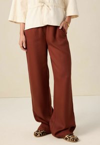 Vrouw in losse bruine broek, crèmekleurige top, gouden armband en ring, en flat shoes met luipaardprint, staand met één hand in haar zak.