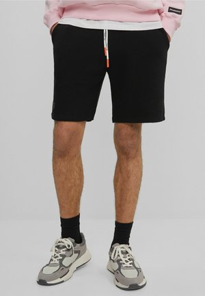 Mand iført sorte shorts, sorte sokker, grå og hvide sneakers samt en lyserød sweatshirt med hænderne i lommerne.