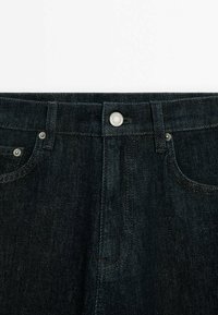 Ceinture de jeans en denim foncé avec bouton métal et passants pour ceinture, montrant les poches avant supérieures et les détails de couture.