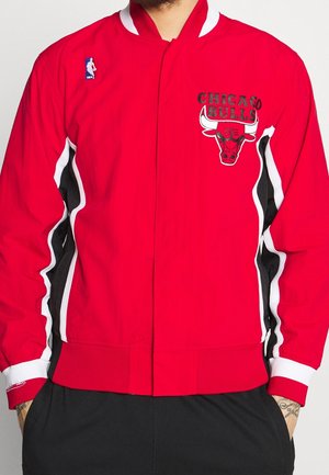 Czerwona kurtka Chicago Bulls z czarnymi i białymi pasami na rękawach oraz patch logo NBA na piersi, noszona z rękami w czarnych kieszeniach spodni.