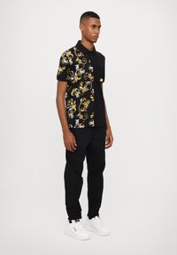 Svart polo skjorte med gul og hvit blomsterdesign på venstre side, ensfarget svart høyre side, korte ermer, parret med svarte joggebukser og hvite sneakers.