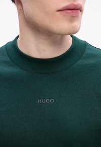 Ciemnozielona bluza z ribbowanym okrągłym dekoltem i wyraźnym szarym logo "HUGO" na piersi; gładka tekstura, dopasowany krój.