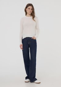 Golf bianco a maniche lunghe abbinato a jeans a palazzo blu scuro. Il modello è in piedi di lato, con le mani nelle tasche. Ai piedi ha delle sneakers bianche.
