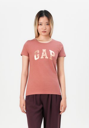 Femme avec des cheveux raides de longueur épaule portant un t-shirt GAP rose poussiéreux et un pantalon violet foncé, se tenant devant un fond blanc.