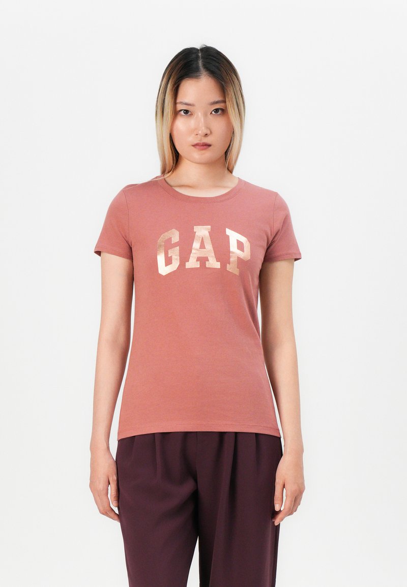 Woman with straight shoulder-length hair wearing a dusty pink GAP t-shirt and dark purple pants, standing against a white background.