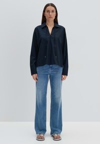 Camicia navy oversize con scollatura a V, maniche lunghe e dettaglio con cordino, abbinata a jeans flare azzurri chiari e sandali flat.