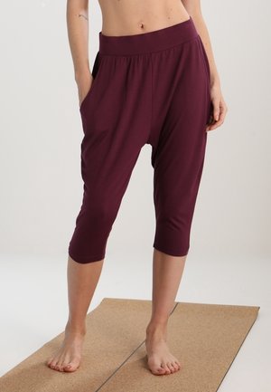 Femme debout pieds nus sur un tapis de yoga portant un pantalon ample bordeaux court avec poches et taille élastique.