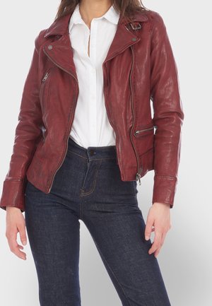 Woman wearing a fitted dark red leather biker jacket over a white button-up shirt and dark blue jeans against a plain background.