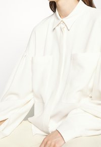 Camicia bianca oversize con colletto, dotata di due tasche frontali e maniche lunghe con polsini, realizzata in un tessuto morbido.
