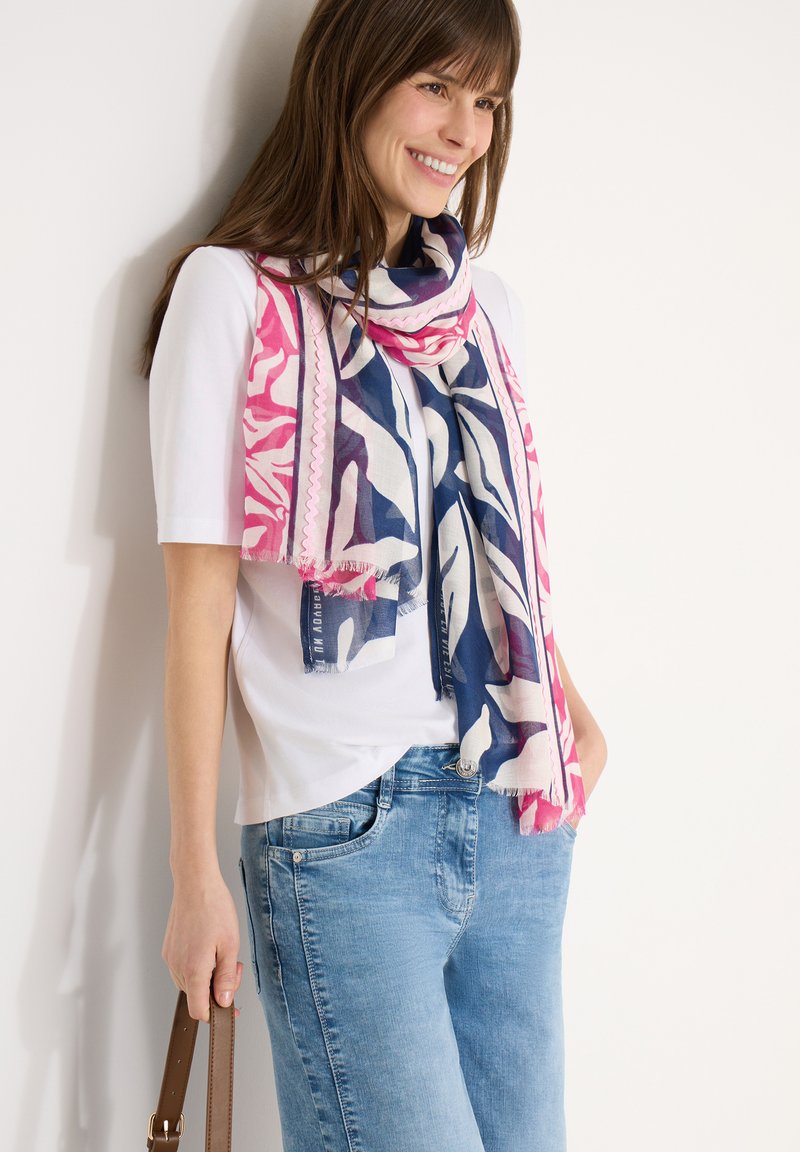 Femme souriante portant une chemise blanche, un jean bleu et une écharpe bleue et rose à motifs, tenant la bandoulière d'un sac à main marron.