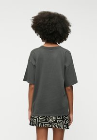 T-shirt oversized gris foncé à manches courtes, col rond et couture visible. Porté sur une jupe noire à motifs avec des designs géométriques blancs.