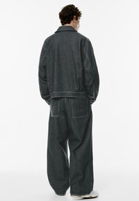 Mørk denimjakke og bukser med brede ben, begge med løs pasform, synlige syninger og lappelommer; bagvinkel mod en ensfarvet baggrund.