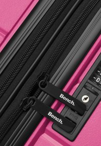 Maleta dura rosa con dos cremalleras negras, que cuenta con tiradores de cremallera de marca y un candado de combinación en el lateral. Superficie texturizada.