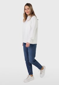 Femme debout de profil portant un sweat-shirt blanc, un jean bleu et des baskets blanches sur un fond clair uni.