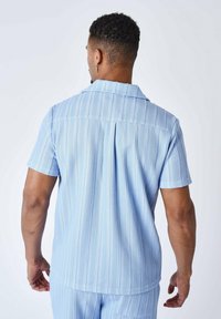 Camicia a maniche corte azzurro chiaro con texture a righe, colletto con bottoni e dettaglio con piega sul retro. Il materiale sembra leggero e informale.