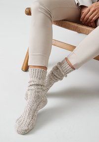 Calcetines de punto beige con una textura acanalada, con un diseño holgado que se extiende por encima del tobillo. Llevados con pantalones ajustados de color claro.