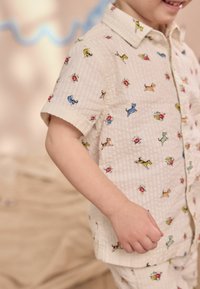 Camicia a maniche corte color crema, con bottoni, a motivo a griglia, caratterizzata da stampe colorate di cani e fiori. Tessuto leggero, vestibilità comoda.