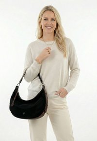 Šviesiaplaukė moteris šypsosi, dėvi kreminės spalvos megztinį ir kelnes, ant peties laiko juodą rankinę su leopardžio rašto apvadu, fone balta spalva.
