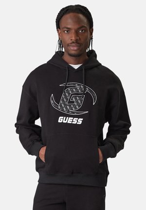 Mann mit geflochtenem Haar trägt schwarzen Guess-Hoodie mit weißem Logo und hat die Hände in den vorderen Taschen vor einfarbigem Hintergrund.