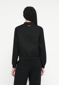 Geacă bomber neagră cu textură fină, manșete și talie ribbed. Prezintă un logo subtil la spate. Stilizată cu pantaloni negri asortați.