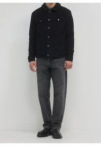 Veste en denim noir avec col en shearling, deux poches avant, fermeture à boutons, portée avec un jean gris foncé et des bottes noires.