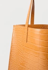 Bež torbica iz umetnega usnja z reliefnim vzorcem krokodila, opremljena z dvema ročajema in strukturirano zasnovo. Brez vidnih kovinskih delov ali dodatkov.