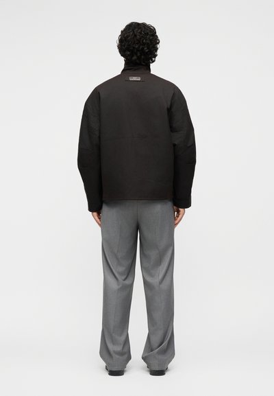 Άτομο με σγουρά μαλλιά, που φοράει μαύρο σακάκι και γκρι παντελόνι, στέκεται γυρισμένο προς τα πίσω σε ένα απλό φωτεινό φόντο.