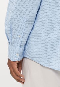 Brazo derecho de una persona vestida con una camisa de manga larga de rayas azul claro y pantalones beige, con la mano relajada a su lado.