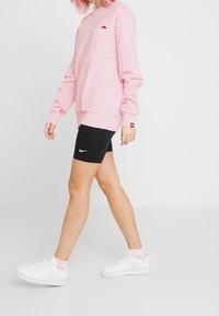 Ljusrosa sweatshirt med ribbstickade muddar, svarta cykelshorts med vitt logo och vita sneakers med rosa strumpor, alla på en enfärgad bakgrund.