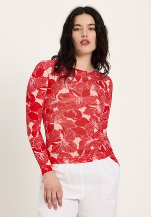 Woman with dark wavy hair wearing a long-sleeve red and pink floral top and white pants, standing with one hand in pocket.