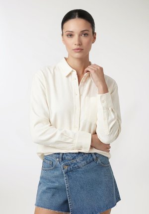 Donna con capelli scuri, camicia crema con bottoni e gonna di jeans asimmetrica, in posa davanti a uno sfondo bianco semplice.