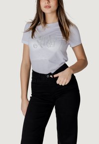 Maglietta bianca con testo glitterato che dice "Scambio di Aeroplani", abbinata a pantaloni neri a vita alta. Tessuto liscio, maniche corte.