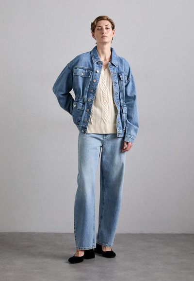 Spijkerjasje met een losse pasvorm, lichtblauwe kleur en grote zakken, gedragen over een crèmekleurige kabeltrui en wijdvallende lichte jeans. Zwarte ballerina's.
