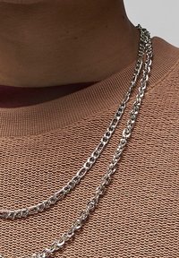 Chaînes métalliques de couleur argentée superposées autour du cou, présentant un design torsadé et des épaisseurs variées, sur un pull marron texturé.