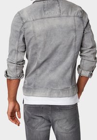 Grå denimjacka med en något blekt finish, klassisk krage och uppvikta ärmar, med sömnadsdetaljer och en subtil logotyp i fållen.