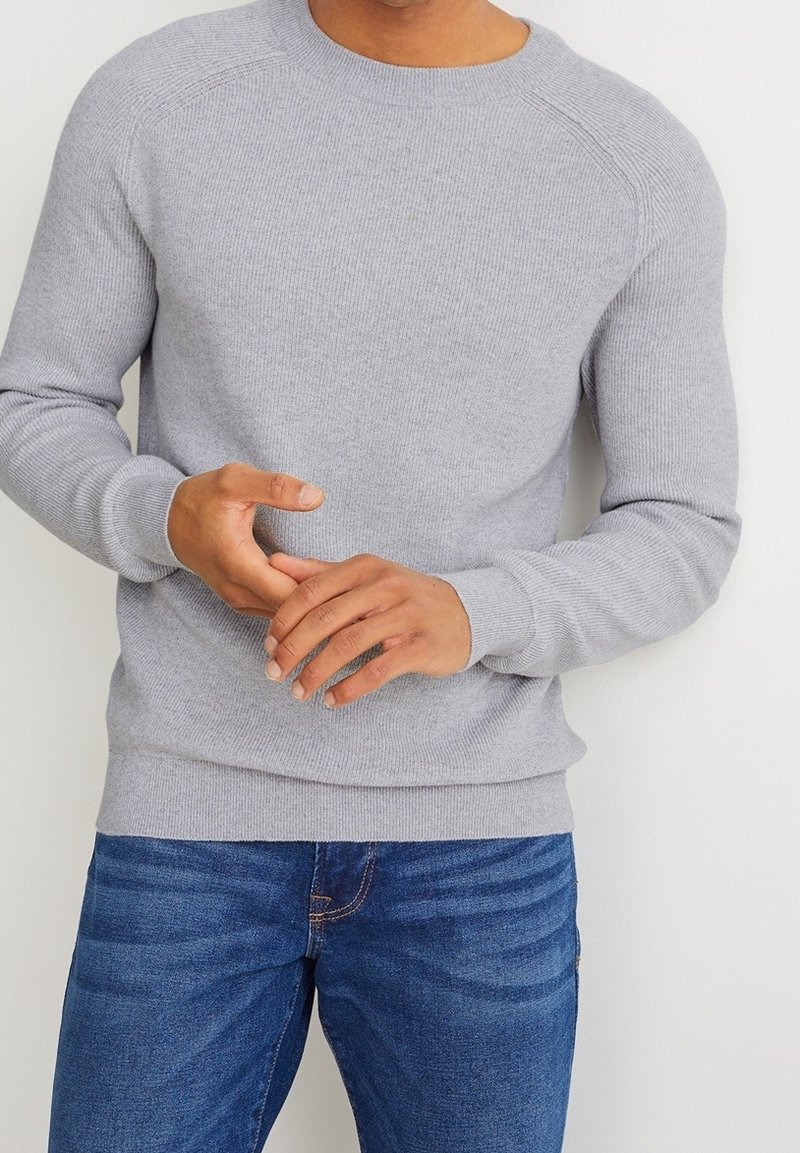 Personne portant un pull en maille côtelée gris clair et un jean bleu, les mains jointes devant, sur un fond blanc uni.