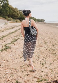 Svart singlet, gjennomsiktig svart skjørt med hvite prikker, og matchende veske med hvite prikker; går på en grusstrand med grøntområde i bakgrunnen.