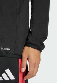 Μαύρο αθλητικό τοπ με μακριά μανίκια, που φέρει το λογότυπο ClimaCool και υφασμάτινη υφή. Το τελείωμα και οι μανσέτες έχουν αντίθετης απόχρωσης φινίρισμα. Ο θρόισμα είναι ορατός.