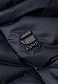 Tessuto trapuntato nero con logo in rilievo con le lettere "RAW" in finitura lucida, che enfatizza una texture liscia e un design moderno.
