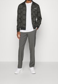 Chaqueta negra con cierre y un patrón ornamental gris, combinada con pantalones de vestir gris claro y zapatillas blancas. Textura de tela suave en toda la prenda.