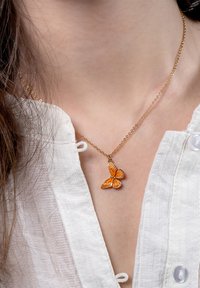 Kuldne kaelakee, millel on oranž liblikapendant keeruliste detailidega. Kandmine valge tekstuuriga kanga topi peal.