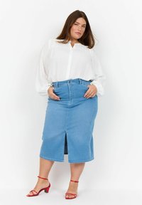 Uslužna bela bluza z dolgimi rokavi in gumbi spredaj, kombinirana z midi krilom iz modrega jeansa, ki ima sprednji razporek, in rdečimi sandali na peto.