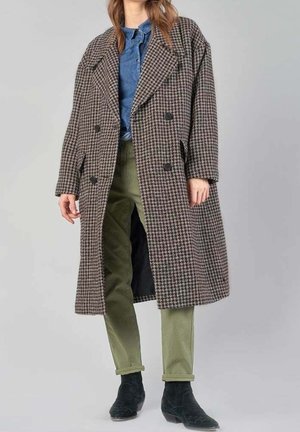 Dubbelzijdige houndstooth jas in bruin en zwart, oversized pasvorm, gedetailleerde revers, gedragen over een blauwe shirt en groene broek, met zwarte enkeltjes.