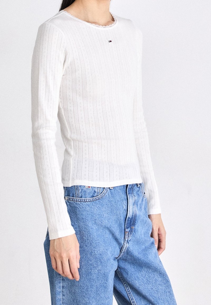 Biała bluzka z długim rękawem o ryflowanej strukturze i koronkowym wykończeniem przy dekolcie, z małym logo. Połączona z niebieskimi dżinsami o wysokim stanie.