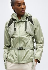 Γυναίκα που φοράει ένα ανοιχτό πράσινο αδιάβροχο μπουφάν και ένα αντίστοιχο σακίδιο πεζοπορίας με ρυθμιζόμενους ιμάντες στο στήθος και στη μέση.