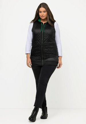 Schwarze gesteppte Weste mit hohem Kragen, grünen Reißverschlussdetails, getragen über einem weißen Langarmoberteil, kombiniert mit schwarzen Jeans und schwarzen Stiefeln.